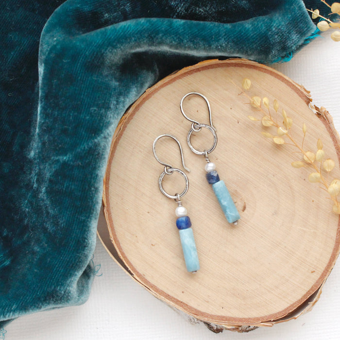 The stacked pearl, kyanite, and larimar hoop earrings styled on wood, blue velvet, and dried grass.