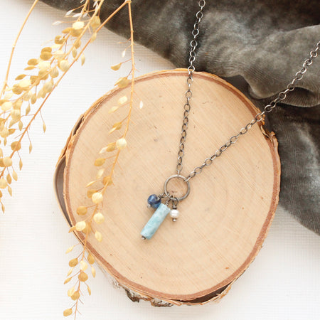 The mirage sydney charm necklace styled on a wooden round, gray velvet, and dried grass.