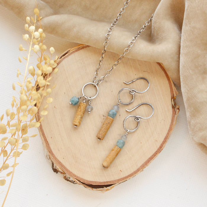 The oasis sydney charm necklace and matching earrings styled on wood with tan velvet and dried grass.
