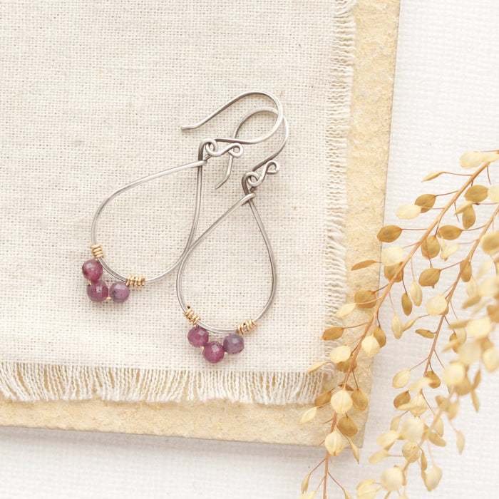 The purple ruby wrapped teardrop hoop earrings styled on limestone, tan linen, and dried grass