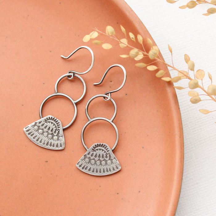 The asmi triangle double loop earrings styled on a red plate with dried grass