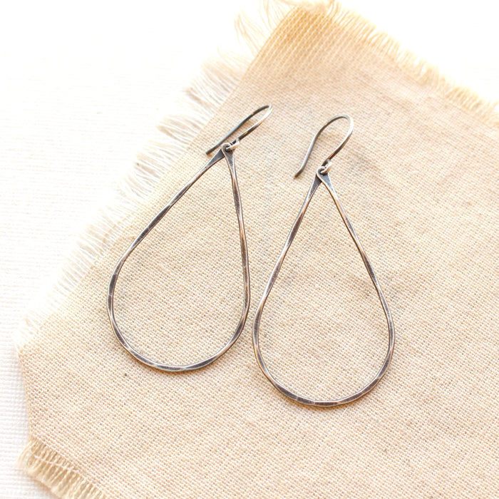 Forged oxidized silver teardrop hoop earrings styled on tan linen.