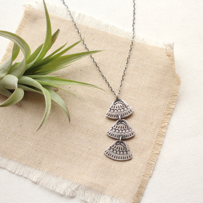 Stamped silver Asmi trio triangle necklace styled on tan linen with an airplant