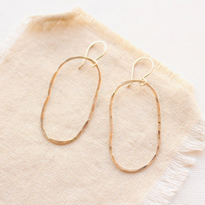 Gold organic shaped oval hoops with a hammered texture styled on tan linen.