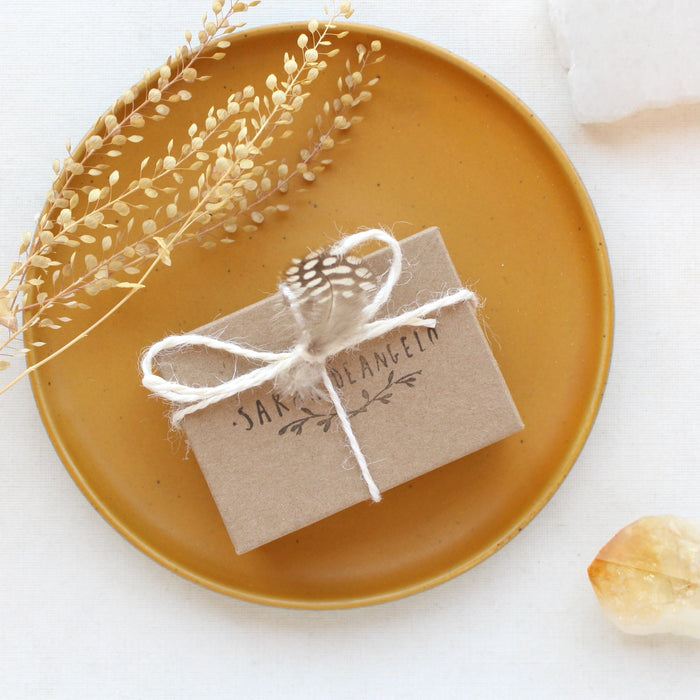 Sarah DeAngleo Jewelry box with signature gift wrap and feather styled on a yellow plate with dried grass and a crystal rock.