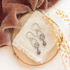 The brown zircon wanderer hoop earrings styled on wood with linen, orange velvet and dried grass