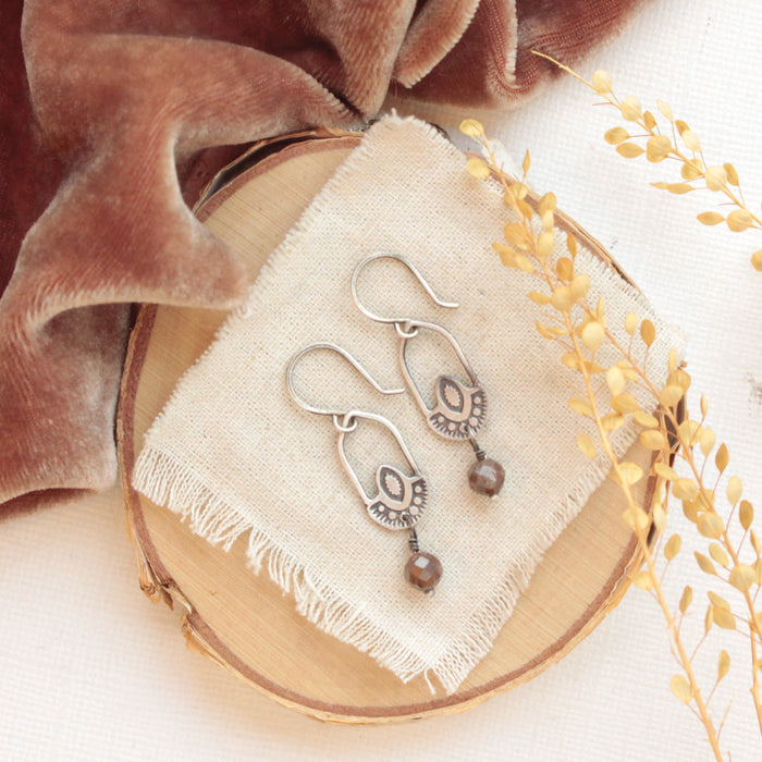 The brown zircon wanderer hoop earrings styled on wood with linen, orange velvet and dried grass