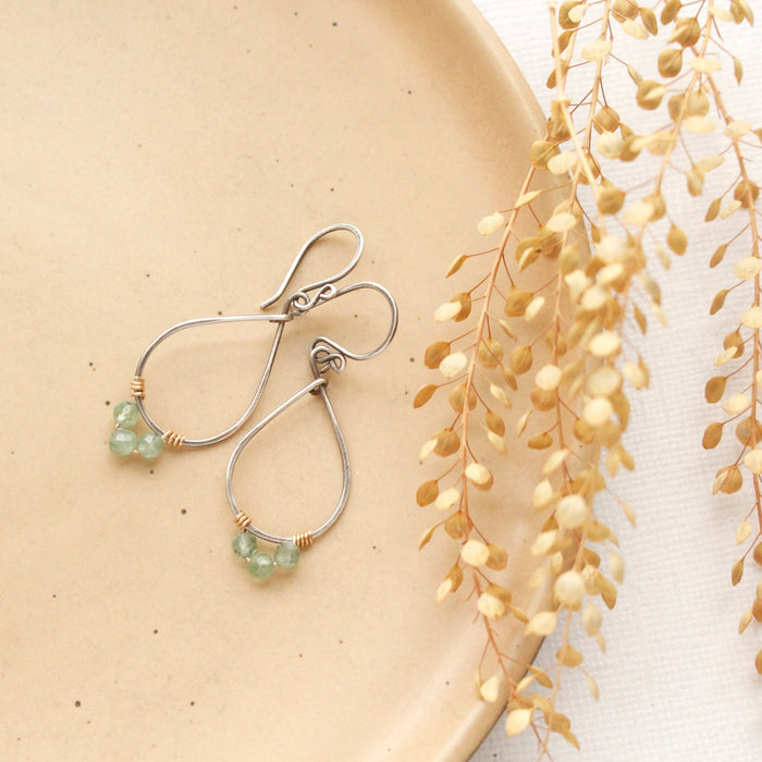 The green apatite wrapped teardrop hoop earrings styled on a tan plate with dried grass