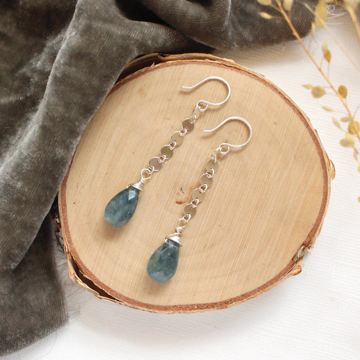 The aquamarine drop and silver disc long earrings styled on wood with gray velvet and dried grass
