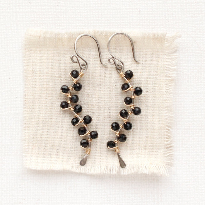 The black tourmaline wrapped mixed metal vine earrings styled on tan linen
