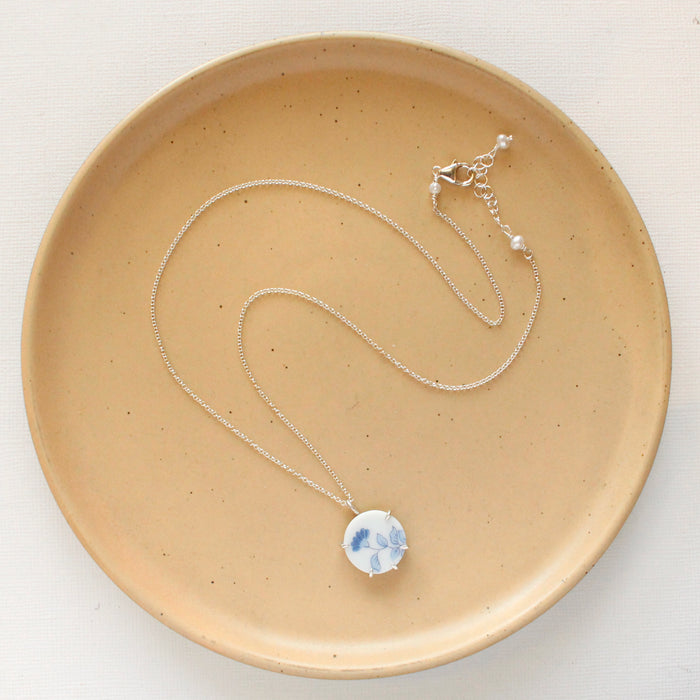 The blue bloom vintage plate necklace styled on a tan plate to show the adjustable lobster clasp closure