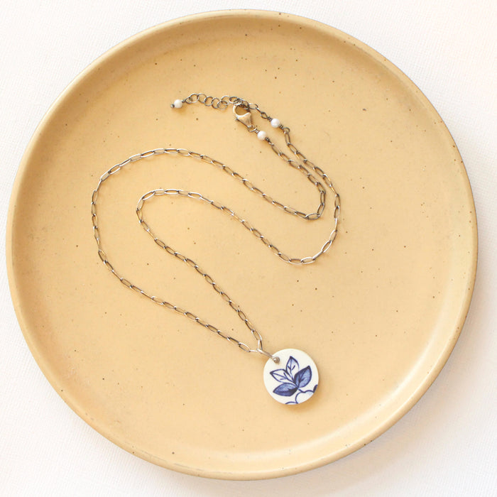 The blue sprig vintage plate necklace styled on a tan plate to show the adjustable lobster clasp closure