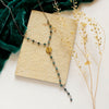 The grandidierite mythos lariat necklace styled on a tan rock with teal velvet and dried grass