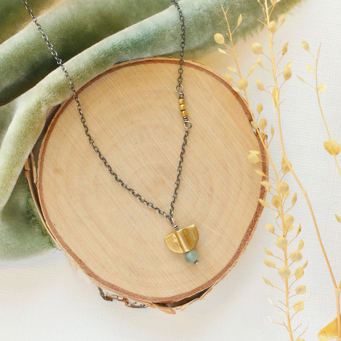 The brass and kyanite mythos necklace styled on wood with green velvet and dried grass