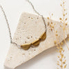 The brass trio mythos necklace styled on a white rock with dried grass