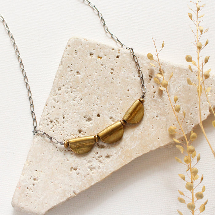 The brass trio mythos necklace styled on a white rock with dried grass