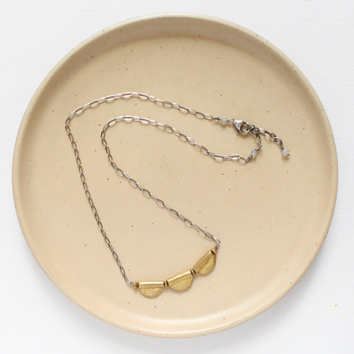 The brass trio mythos necklace styled on a tan plate to show the adjustable lobster clasp closure