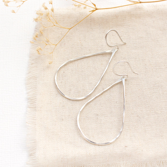 The bright silver forged teardrop hoop earrings styled on tan linen with dried grass
