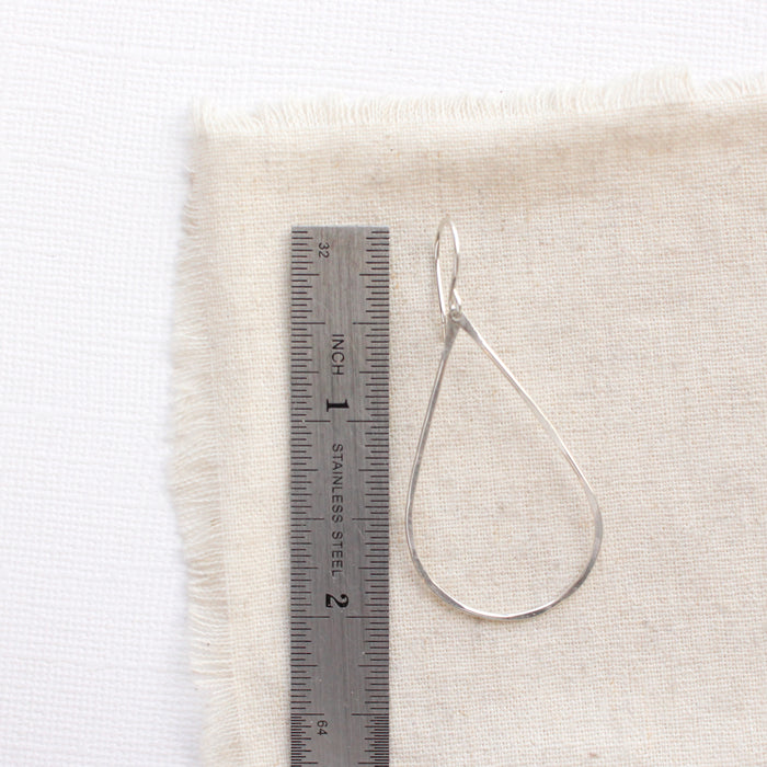 A bright silver forged teardrop hoop earring next to a ruler for size reference