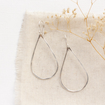 The bright silver forged teardrop hoop earrings styled on tan linen with dried grass