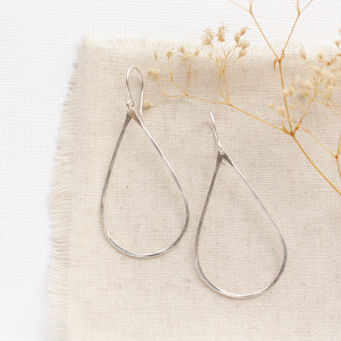 The bright silver forged teardrop hoop earrings styled on tan linen with dried grass