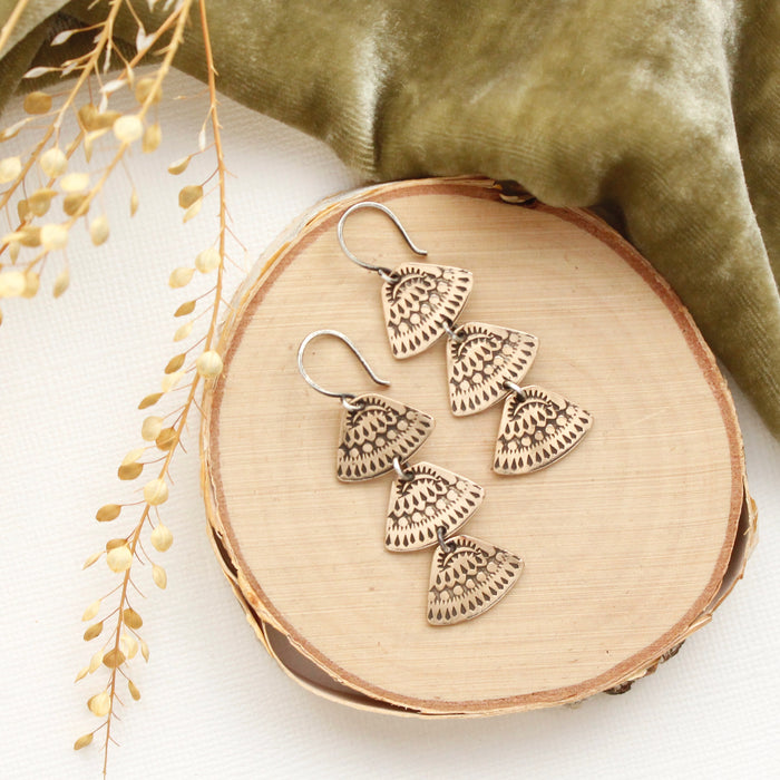 The bronze asmi trio triangle earrings styled on wood with green velvet and dried grass