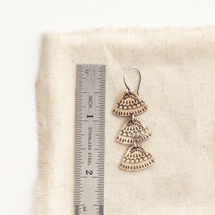 A bronze asmi trio triangle earring next to a ruler for size reference