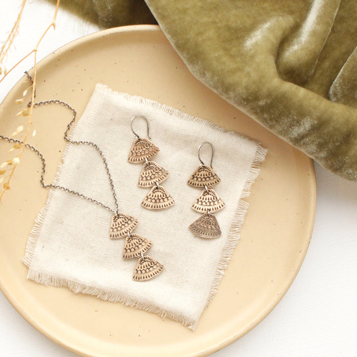 The bronze asmi trio triangle necklace styled on a tan plate with the matching earrings