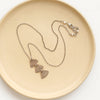 The bronze asmi trio triangle necklace styled on a tan plate to show the adjustable lobster clasp closure