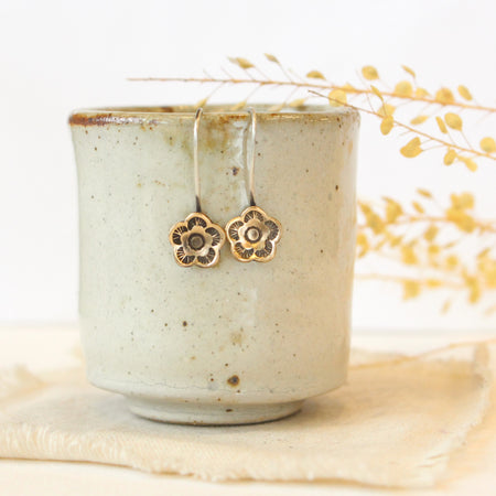 The bronze mini cactus flower lobe hugger earrings styled on a ceramic cup with linen and dried grass