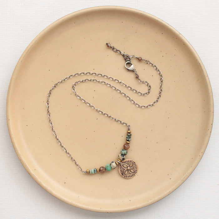 The bronze wanderer lona necklace styled on a tan plate to show the adjustable lobster clasp closure