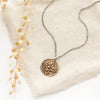 The bronze wanderer coin necklace styled on tan linen with dried grass