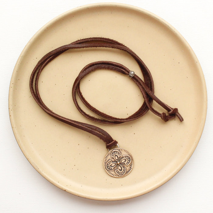 The bronze wanderer medallion leather necklace styled on a tan plate to show the adjustable silver slider