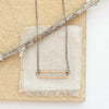 The bronze open bar with silver chain necklace styled on a tan rock with tan linen and a wooden stick