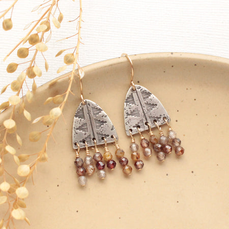 The brown zircon pakal arch fringe earrings styled on a tan plate with dried grass