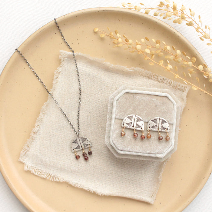 The brown zircon pakal arch post earrings styled with the matching necklace on a tan plate with linen and dried grass