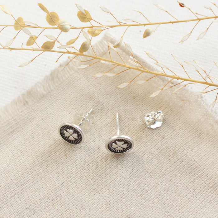 The stamped silver clover post earrings styled on tan linen with dried grass