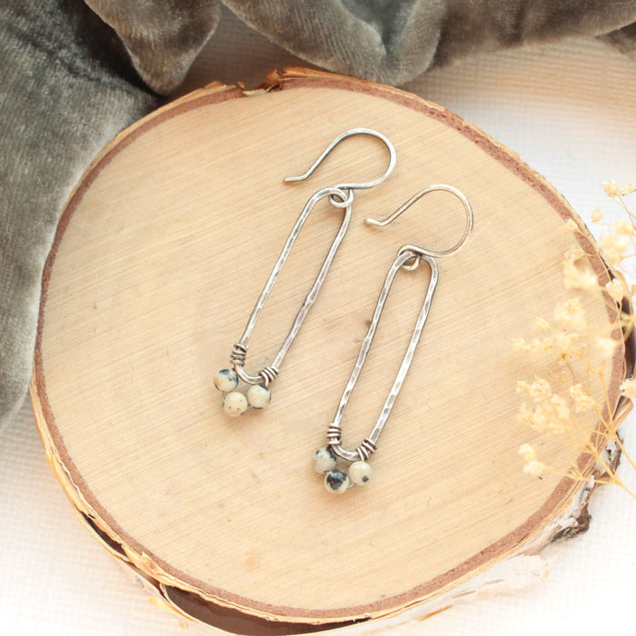 The dalmatian jasper long forged hoop earrings styled on wood with gray velvet and dried grass