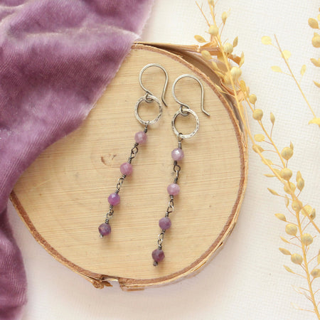 The flowing ruby mini hoop earrings styled on wood with purple velvet and dried grass
