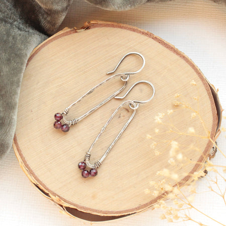 The garnet wrapped long forged hoop earrings styled on wood with gray velvet and dried grass