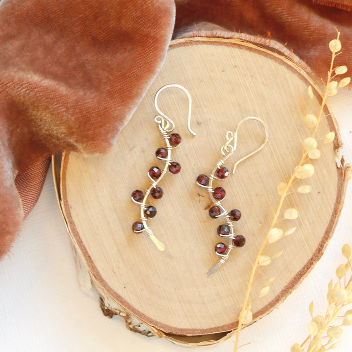 The garnet wrapped silver vine earrings styled on wood with orange velvet and dried grass