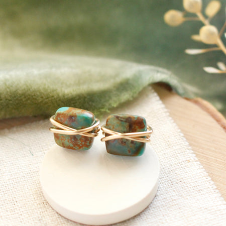 The gold wrapped turquoise square post earrings styled on linen with green velvet and dried grass