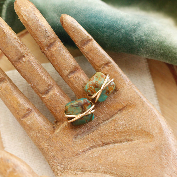 The gold wrapped turquoise post earrings styled on a wooden hand with green velvet and linen