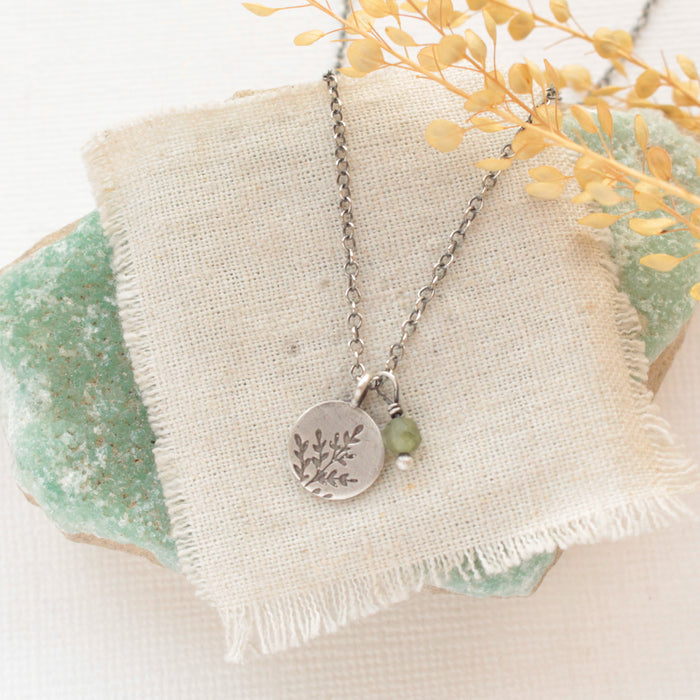 The green kyanite and sprout charm necklace styled on a green rock with tan linen and dried grass
