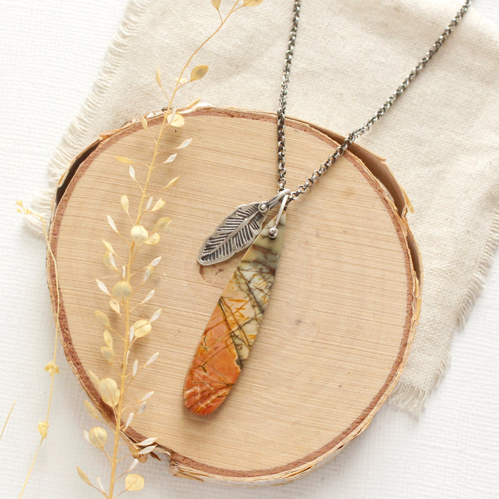 The red creek jasper and feather necklace styled on wood with dried grass and tan linen