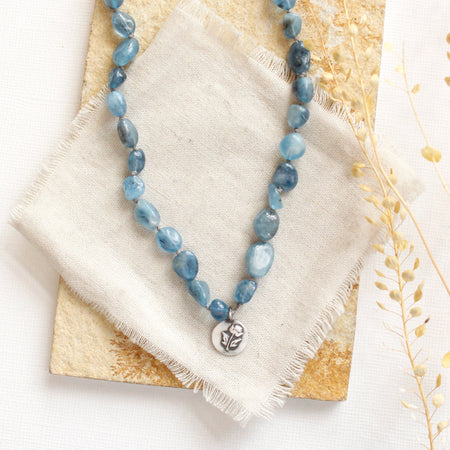 The knotted aqumarine rose necklace styled on a tan rock with linen and dried grass