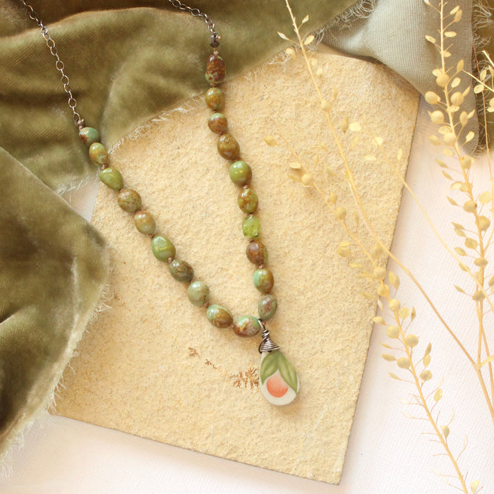 The knotted elisa turquoise, peridot, and peach blossom necklace styled on a tan plate with green velvet and dried grass