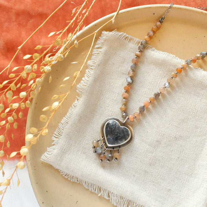 The knotted moonstone heart necklace styled on a tan plate with linen, orange velvet, and dried grass