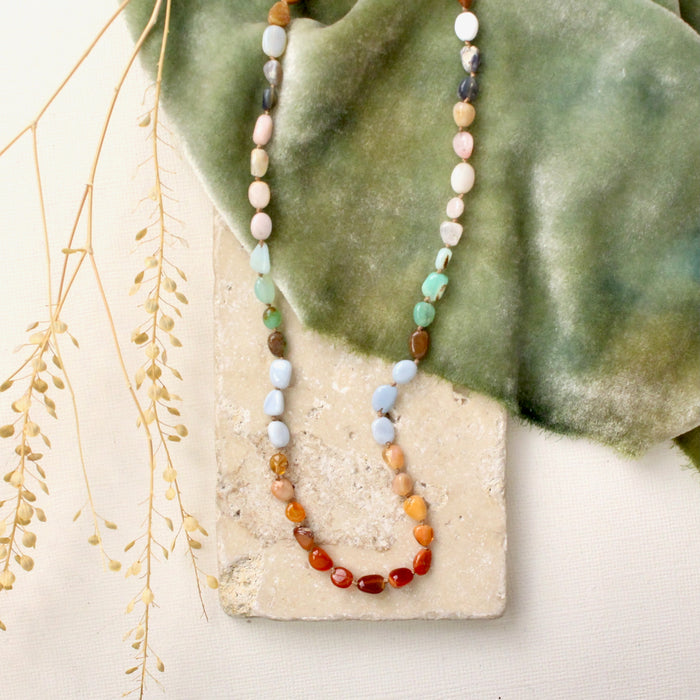 The knotted rainbow opal necklace styled on a tan rock with green velvet and dried grass