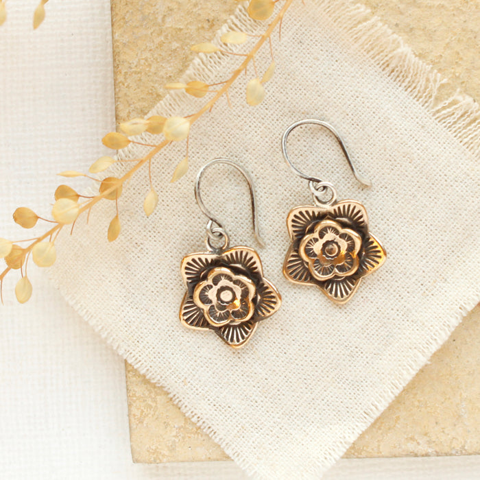 The bronze layered cactus flower earrings styled on a tan rock with linen and dried grass